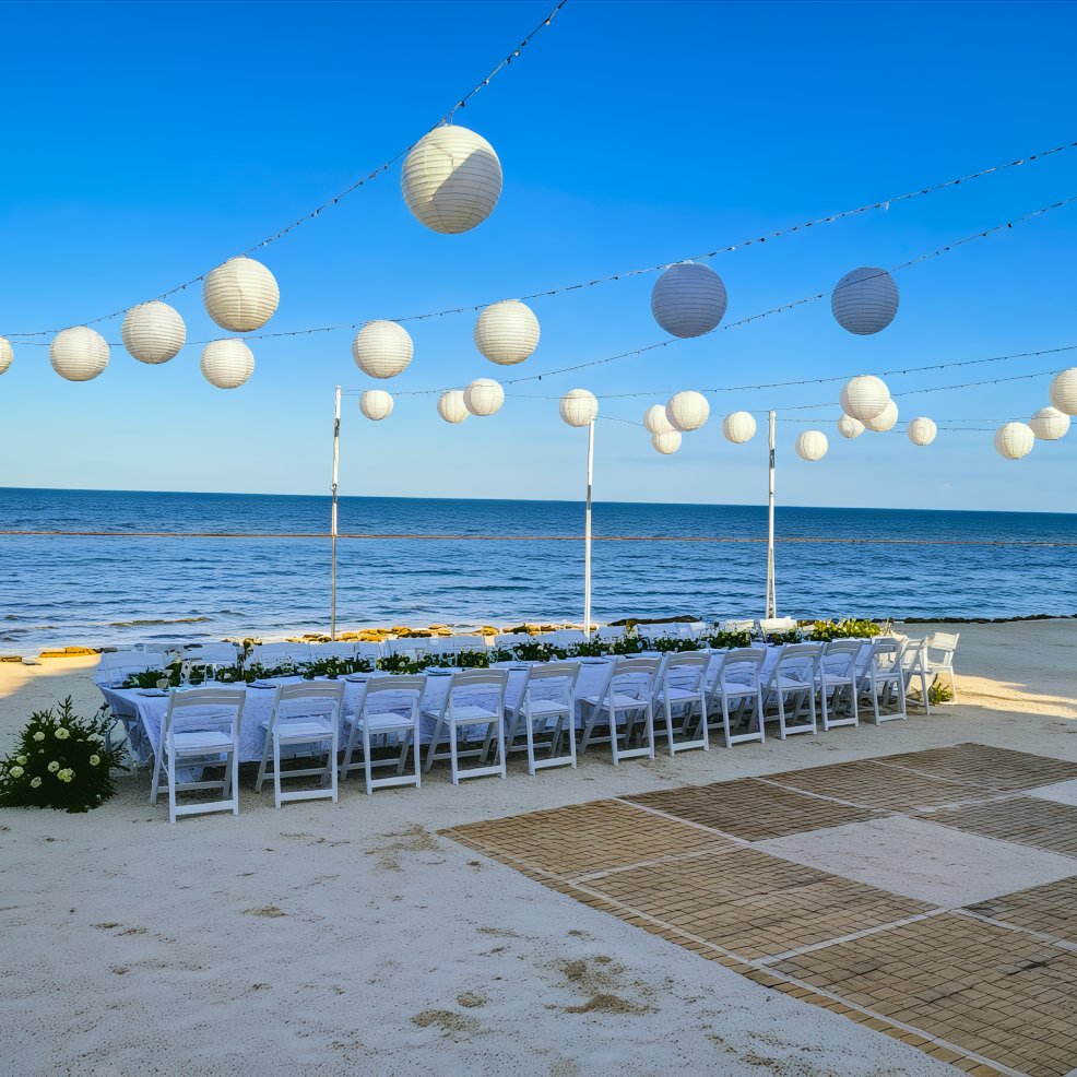 A stunning destination wedding setup on the beachfront features elegant white chairs and tables adorned with paper lanterns, all set against a breathtaking sea view.