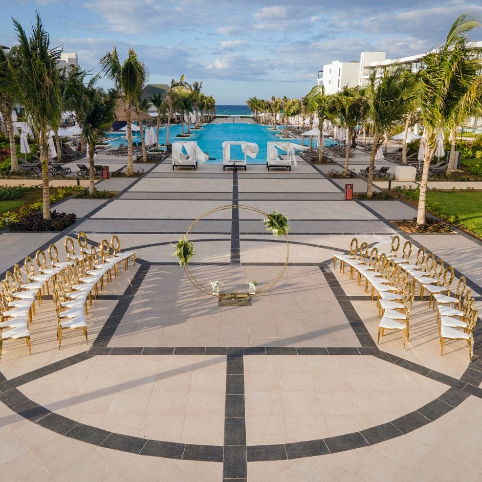 Destination wedding ceremony arranged by a pool, featuring chairs, palm trees, and a circular arch.