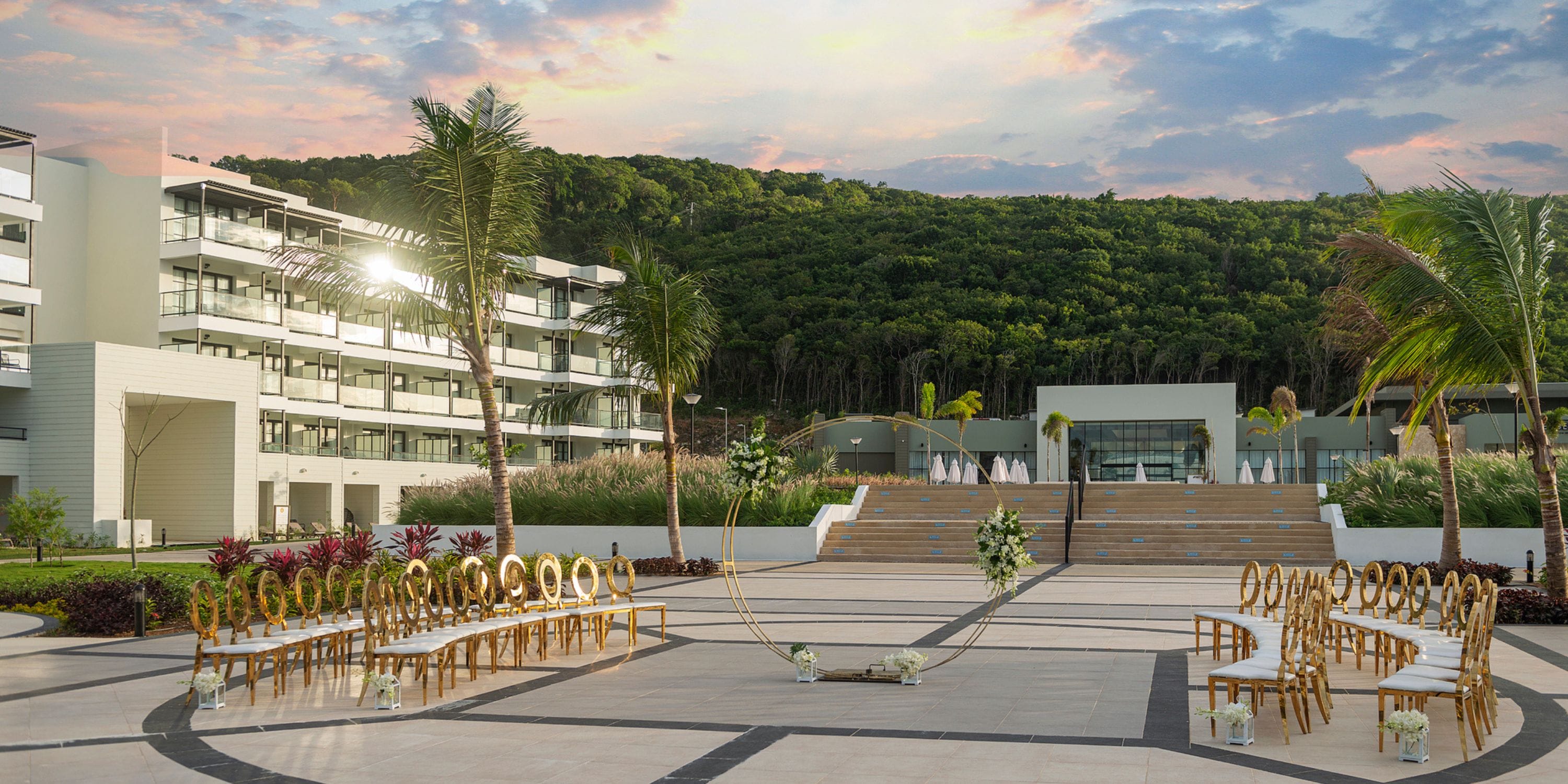 Destination wedding ceremony arranged outdoors with curved seating, modern buildings, and green hill.