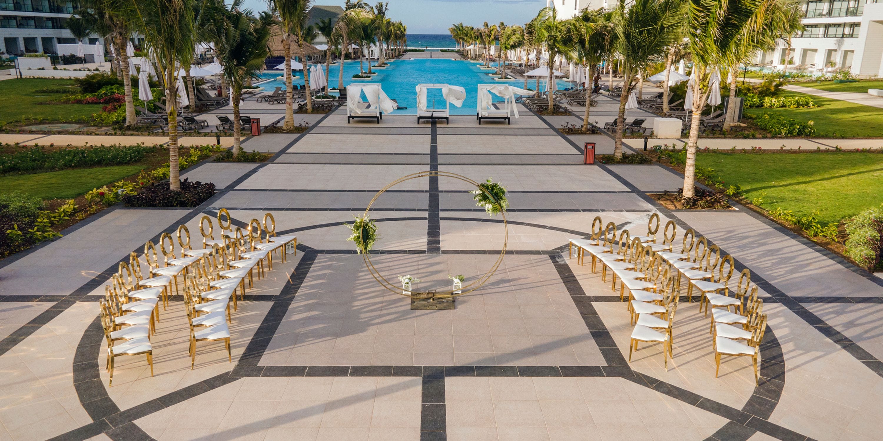 Destination wedding scene featuring gold chairs in a semicircle around a circular arch beside a pool.