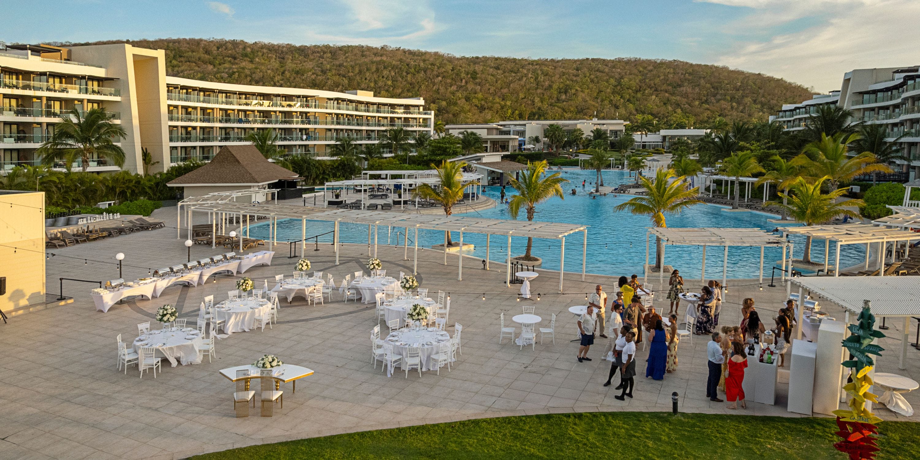 Destination wedding setup with round tables arranged by a large resort pool and contemporary hotel buildings.