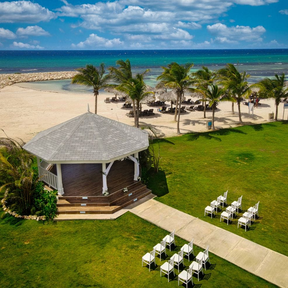 A white gazebo and chairs on a lawn, set for a destination wedding by a beach with palm trees and ocean.