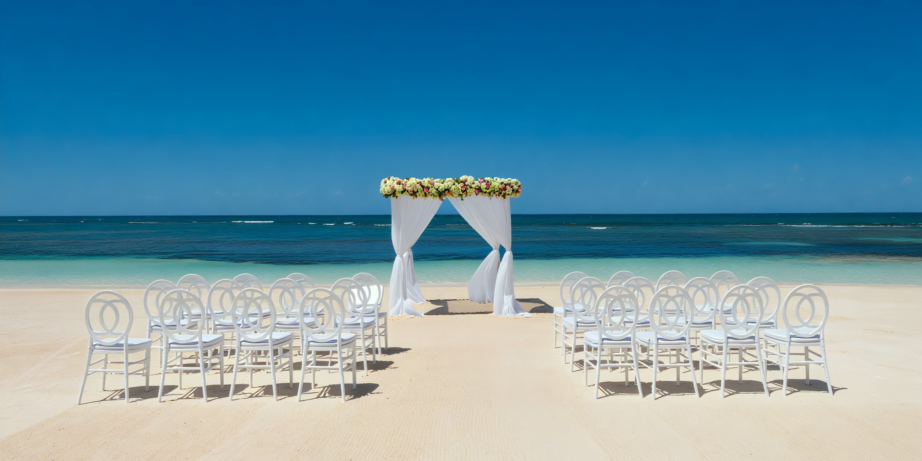 A stunning destination wedding setup featuring white chairs and a floral arch, all beautifully arranged to face the ocean.