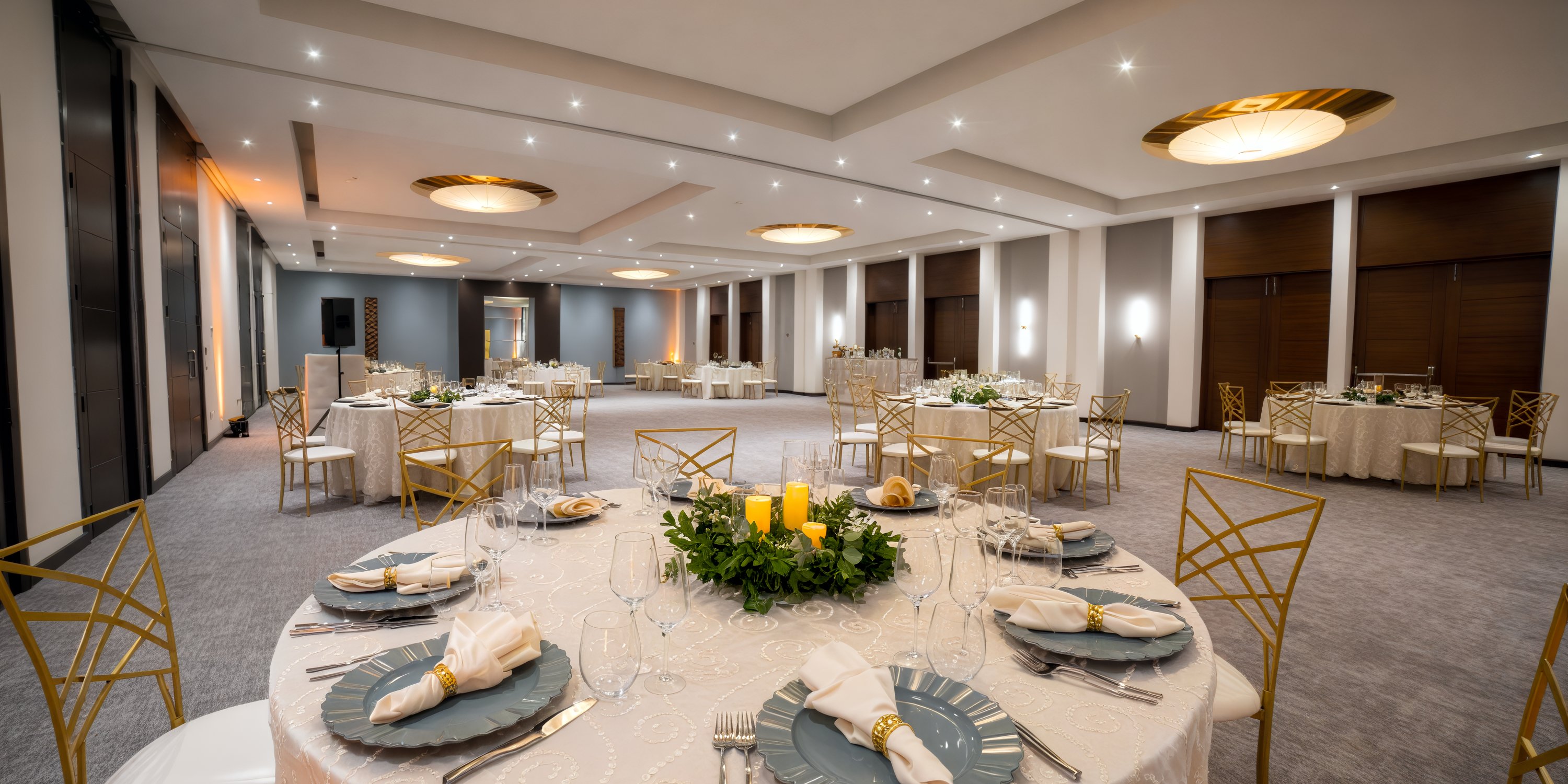 A sophisticated banquet hall beautifully arranged for a destination wedding, featuring round tables adorned with white linens and complemented by elegant gold chairs, sets the perfect ambiance for the event.