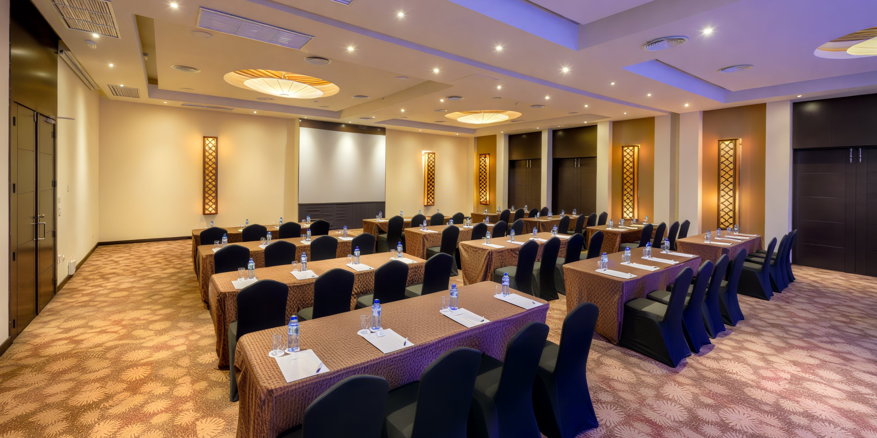 A conference room arranged with rows of tables and chairs, complemented by a projector screen, perfect for hosting a destination wedding.