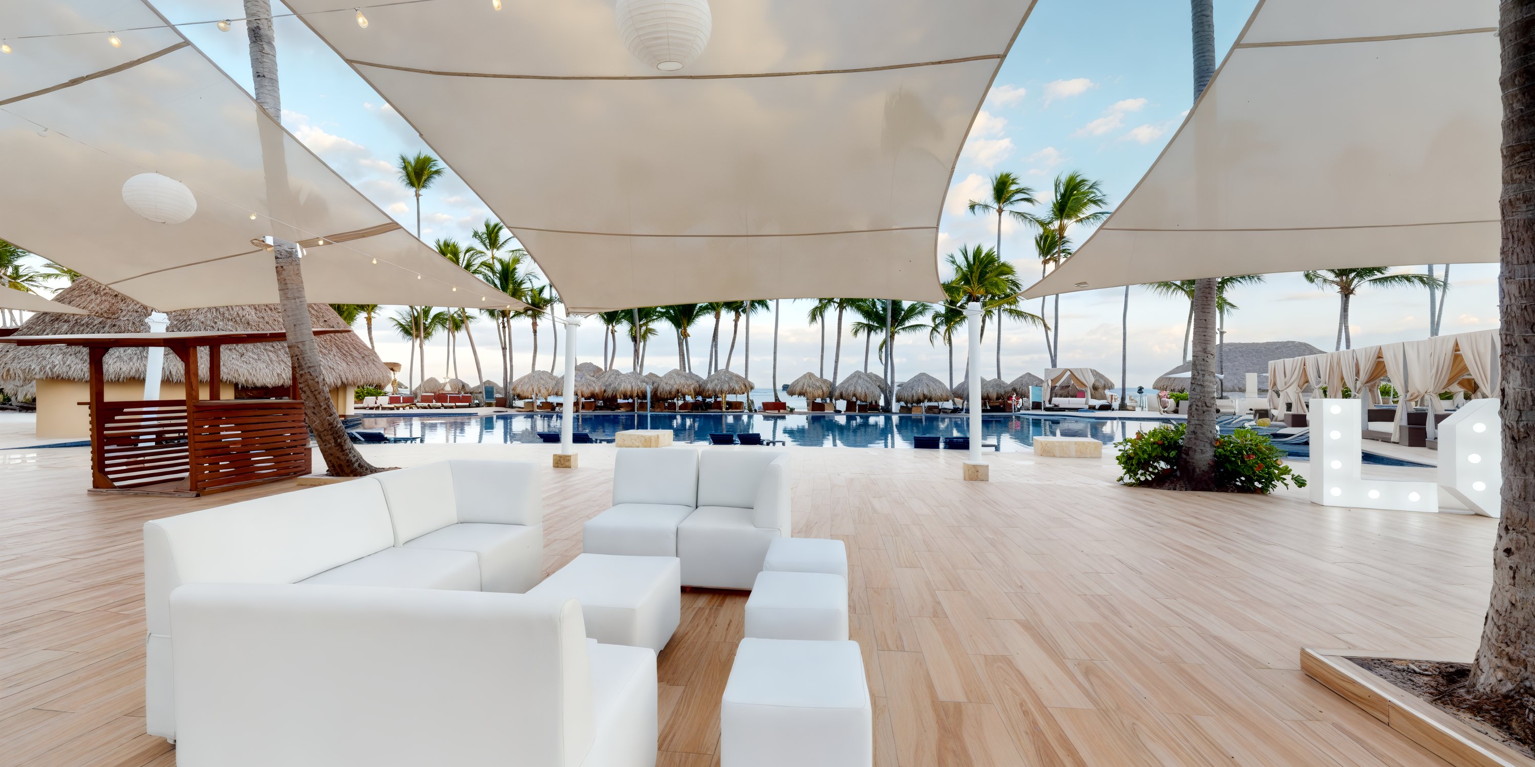White outdoor furniture sits elegantly under canopies by the pool, creating a picturesque setting ideal for a destination wedding, with palm trees and charming huts enhancing the idyllic backdrop.