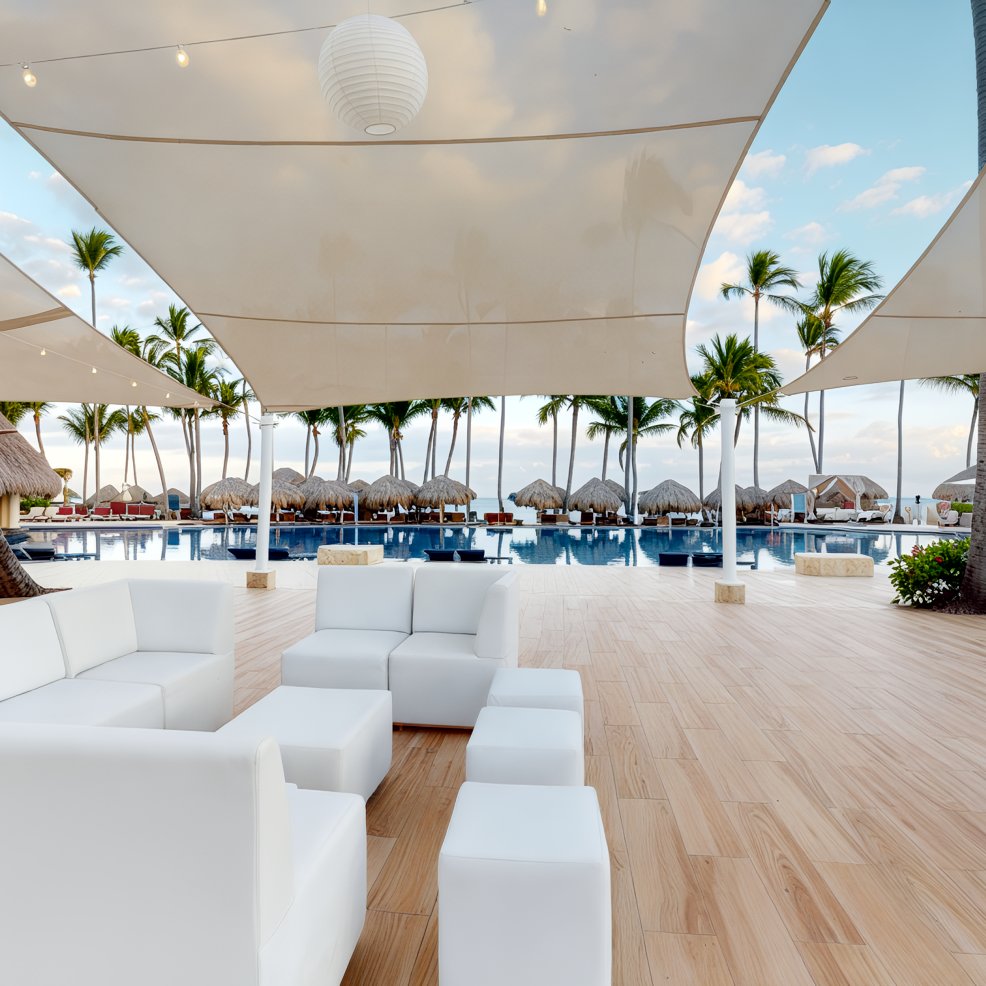 An outdoor patio with elegant white furniture set near a sparkling pool offers a picturesque setting, ideal for a destination wedding, with majestic palm trees creating a stunning backdrop.