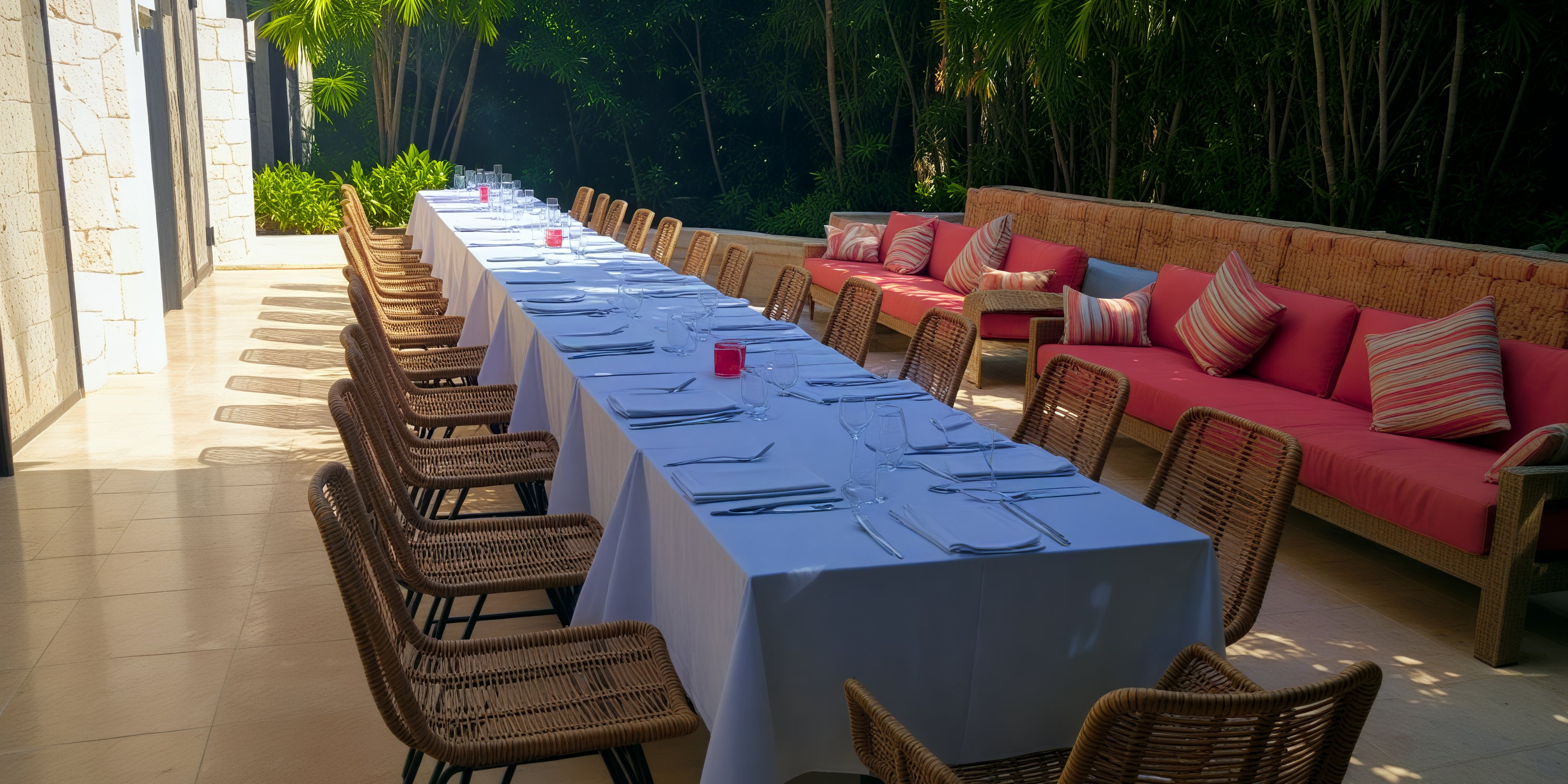 An outdoor dining table set elegantly for a meal, featuring wicker chairs and benches adorned with red cushions, perfect for a destination wedding reception.