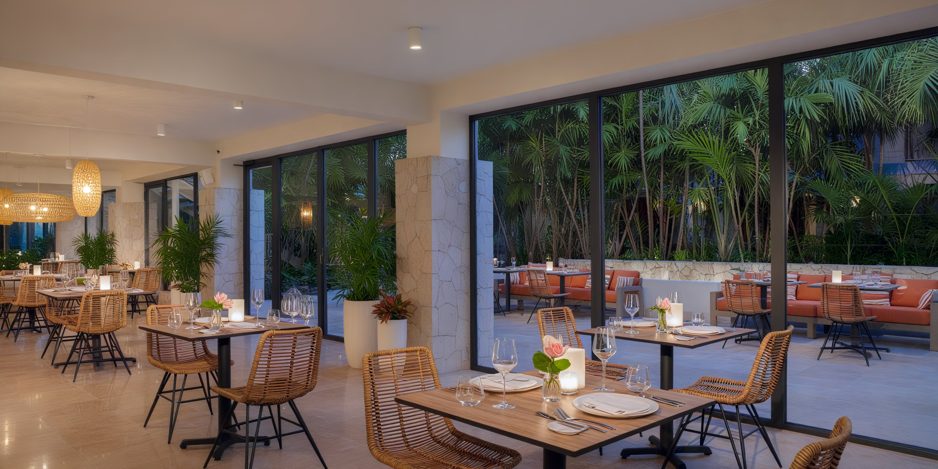 A contemporary restaurant featuring wicker chairs and elegantly set tables, with expansive windows offering views of lush greenery — an ideal setting for a wedding or destination wedding.
