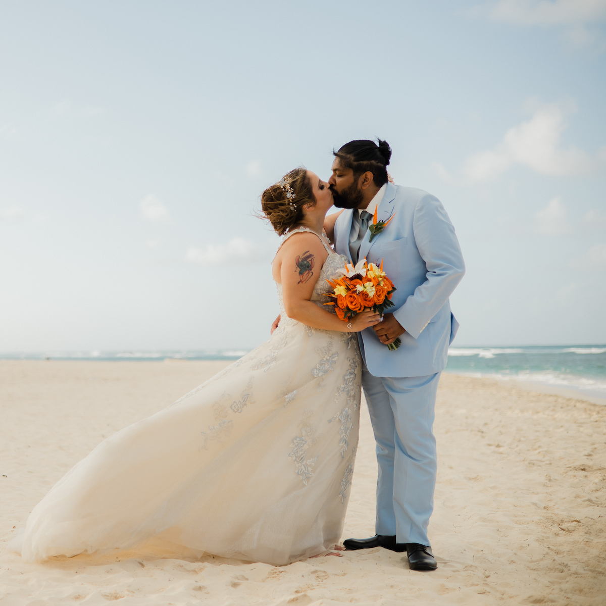 Dressed in their wedding attire and holding a vibrant orange bouquet, Albrianna and Nicholas share a romantic kiss on the beach, creating a picturesque scene from their perfect destination wedding.
