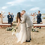 A couple shares a kiss at their beachside destination wedding, surrounded by guests who cheer them on in the background.
