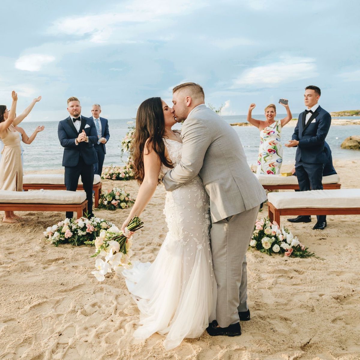 A couple shares a kiss at their beachside destination wedding, surrounded by guests who cheer them on in the background.