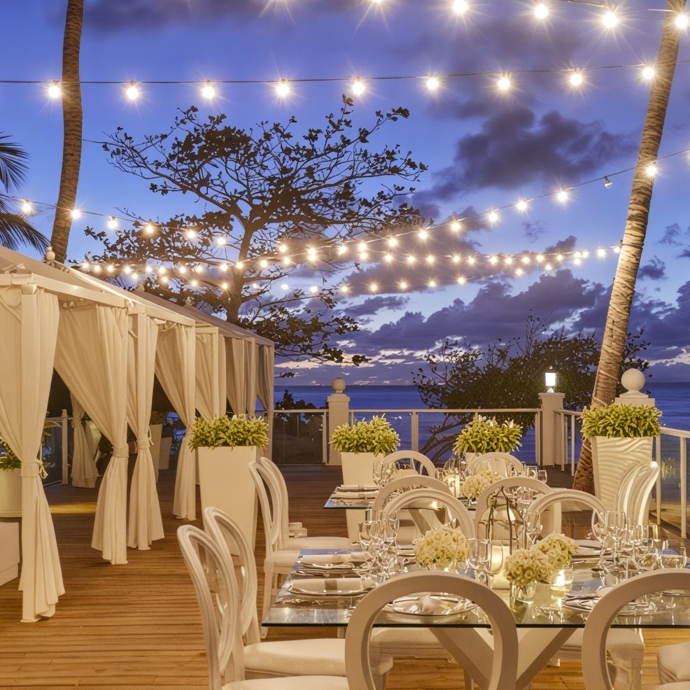 A destination wedding dining arrangement is beautifully set up under string lights at dusk, offering a breathtaking ocean view in the background.