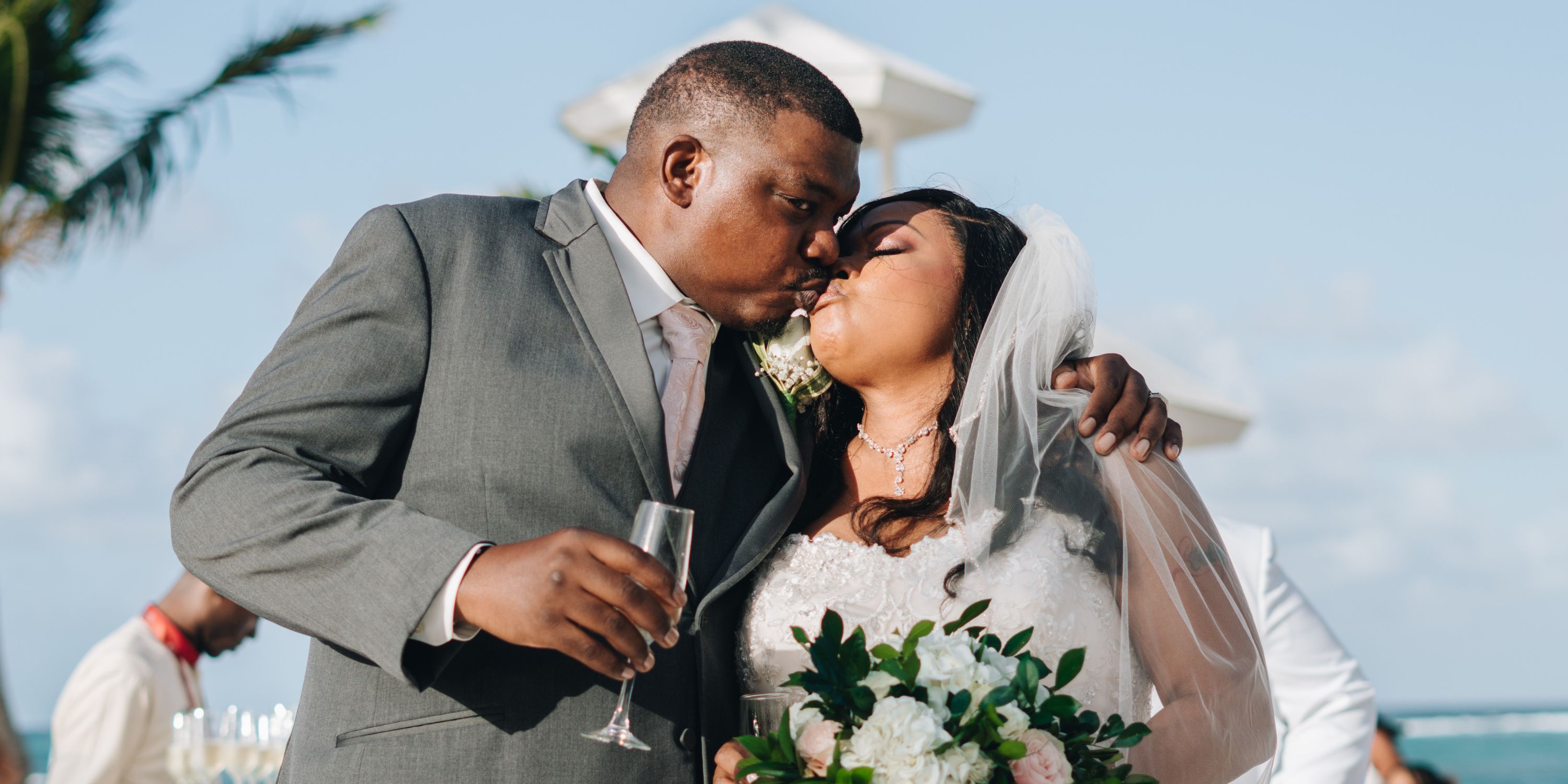 Cheryl and Daryl: The bride and groom share a kiss outdoors at their destination wedding, holding a bouquet and a champagne glass.