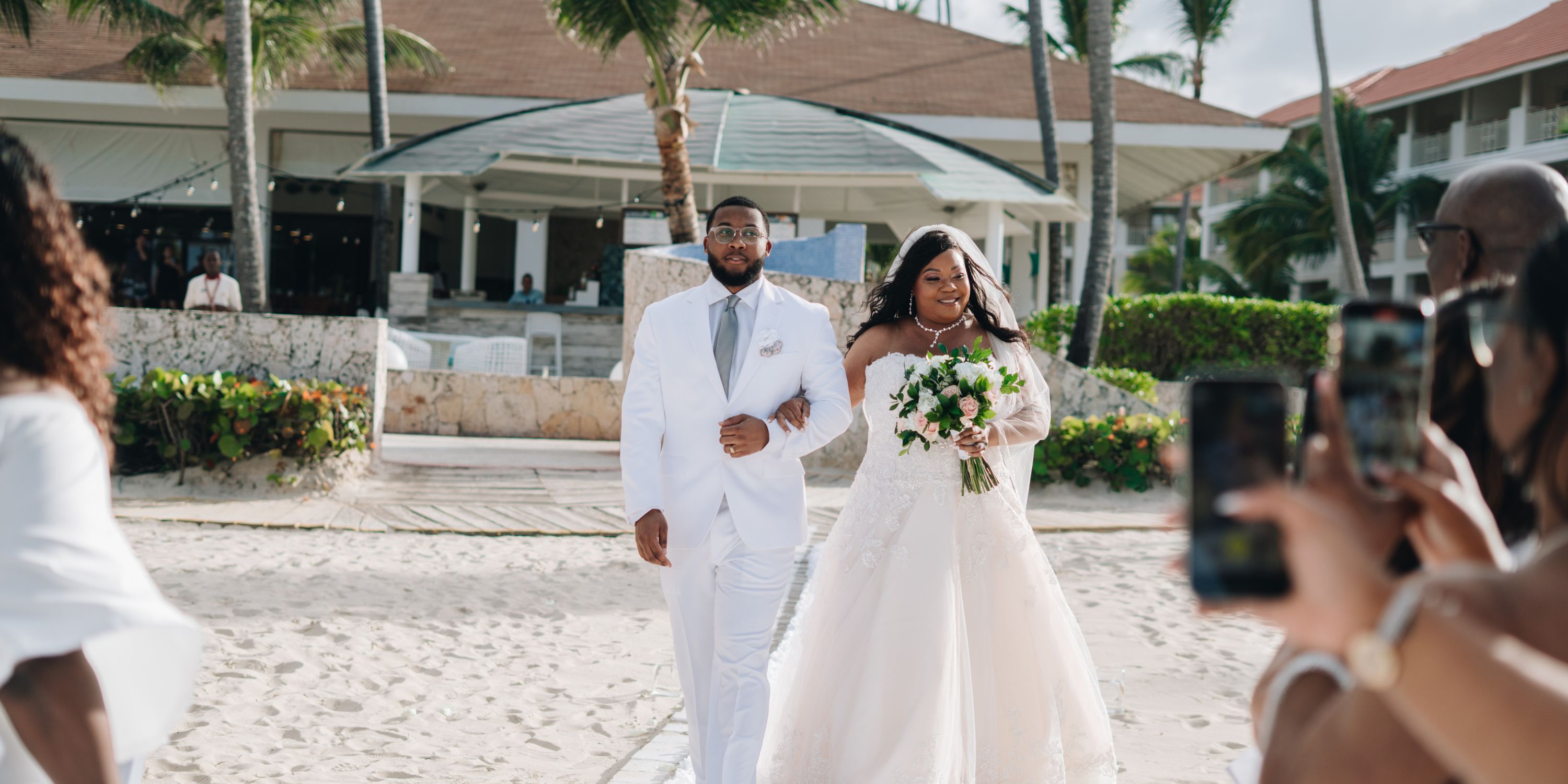 Cathy and Berwick stroll along the sandy beach, enjoying their destination wedding as guests snap photos outside the resort.