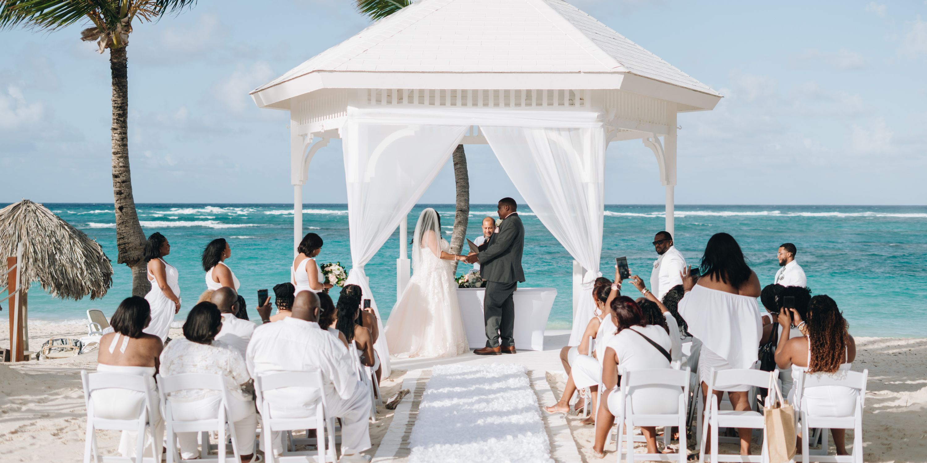 Cathy and Berwick stand beneath a charming gazebo during their destination wedding on the beach, while their guests are seated close by to witness the ceremony.