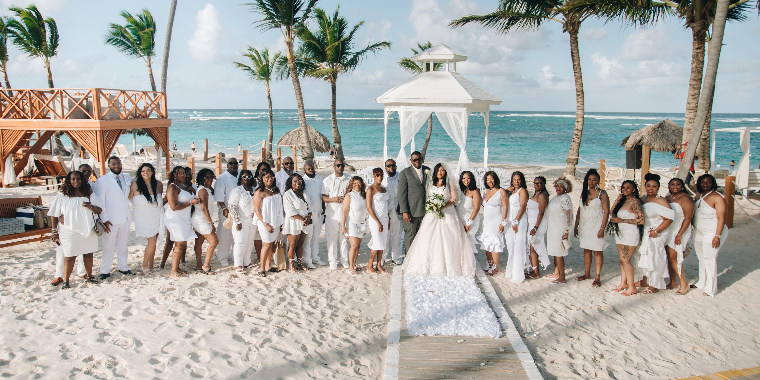 Cathy and Berwick pose with their wedding party on a sandy beach by the ocean, capturing a beautiful moment from their destination wedding.