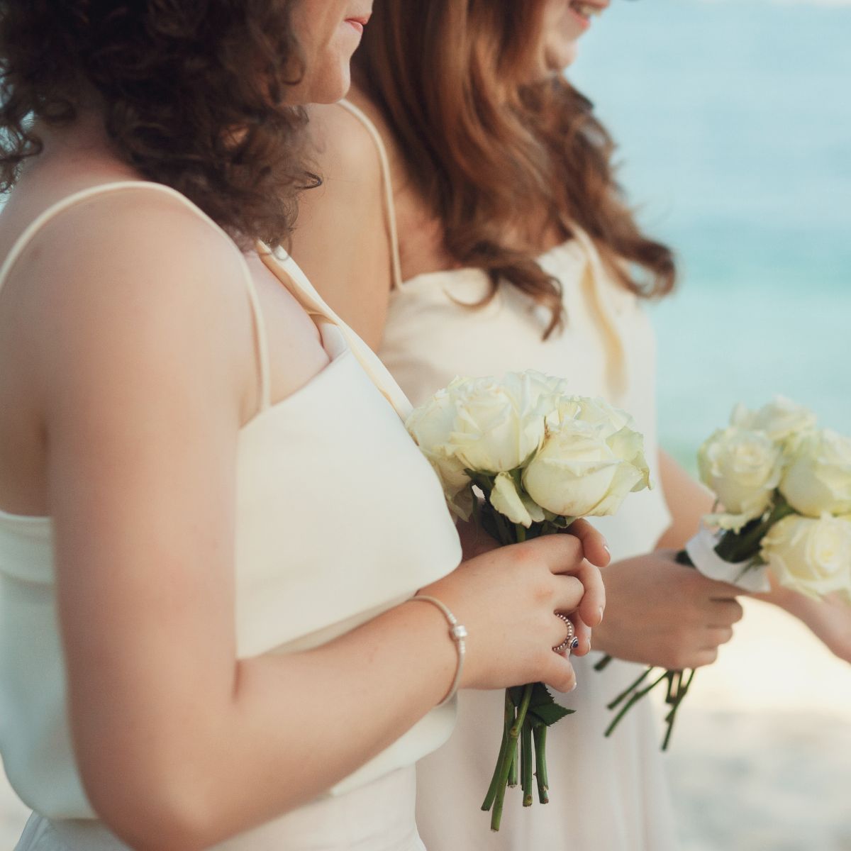 At a destination wedding, two women accessorize their white dresses with petite bouquets of roses at an outdoor celebration.