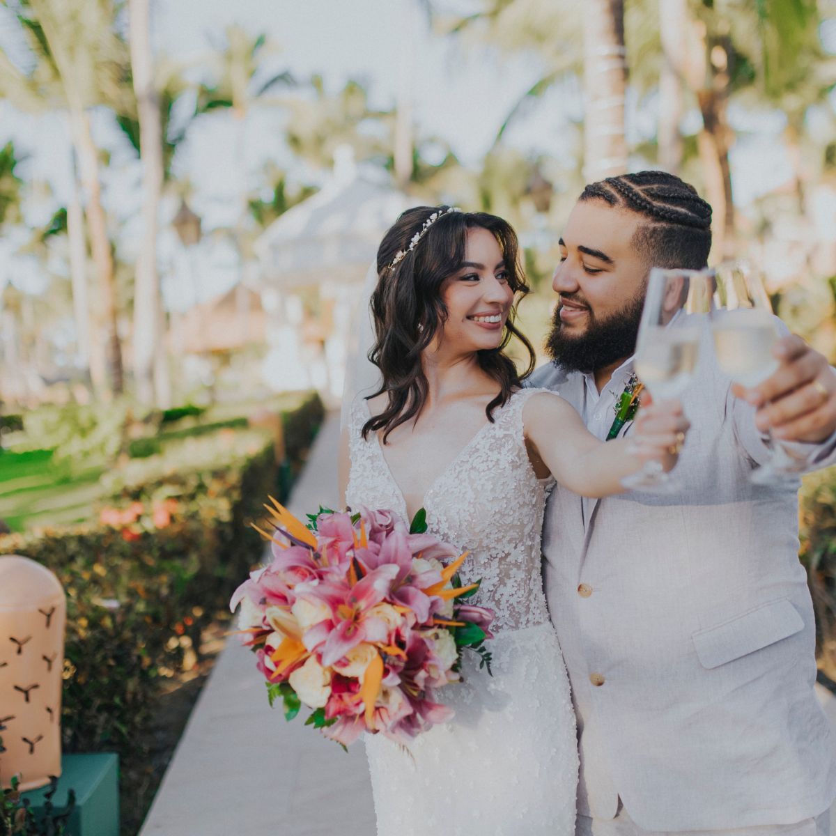 The bride and groom share a joyful smile as they toast with champagne outdoors at their destination wedding, holding a beautiful bouquet of flowers.
