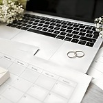 Two wedding rings rest on a laptop alongside flowers and a calendar, all arranged neatly on a desk—setting the scene for planning a destination wedding.