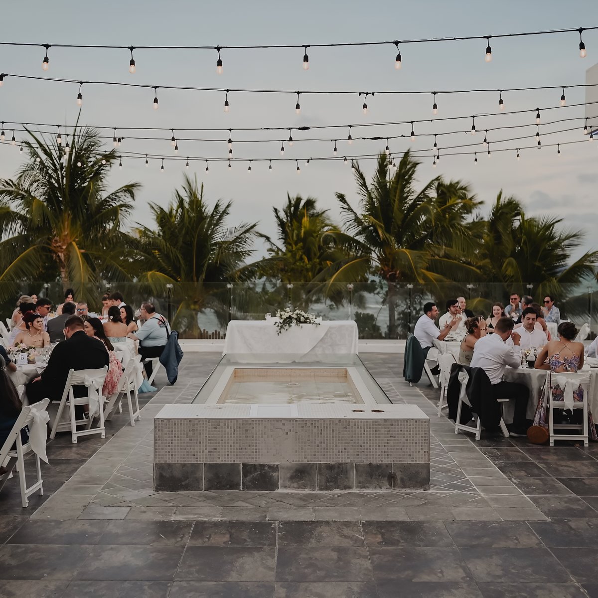 A beautiful destination wedding with guests seated at elegant tables near a sparkling fountain, twinkling string lights overhead, and majestic palm trees in the background.