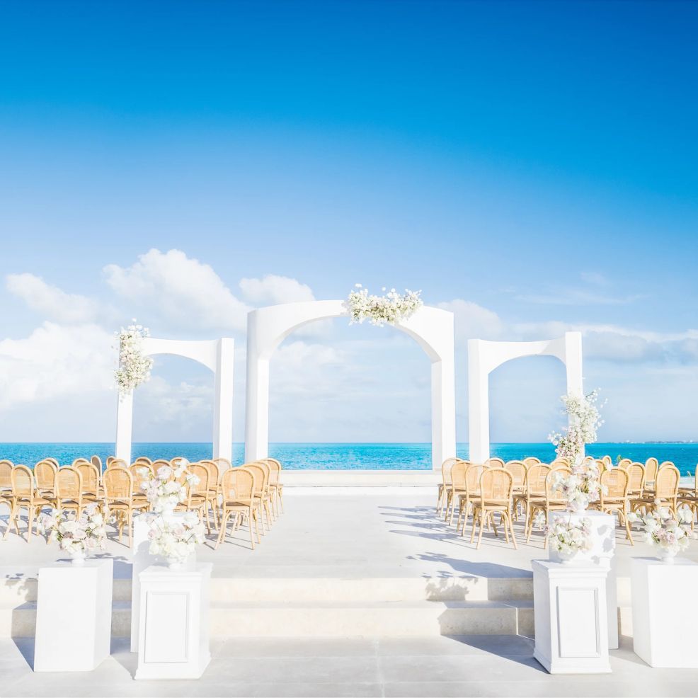 Destination wedding setup featuring white arches, rows of chairs, and floral arrangements by the ocean.