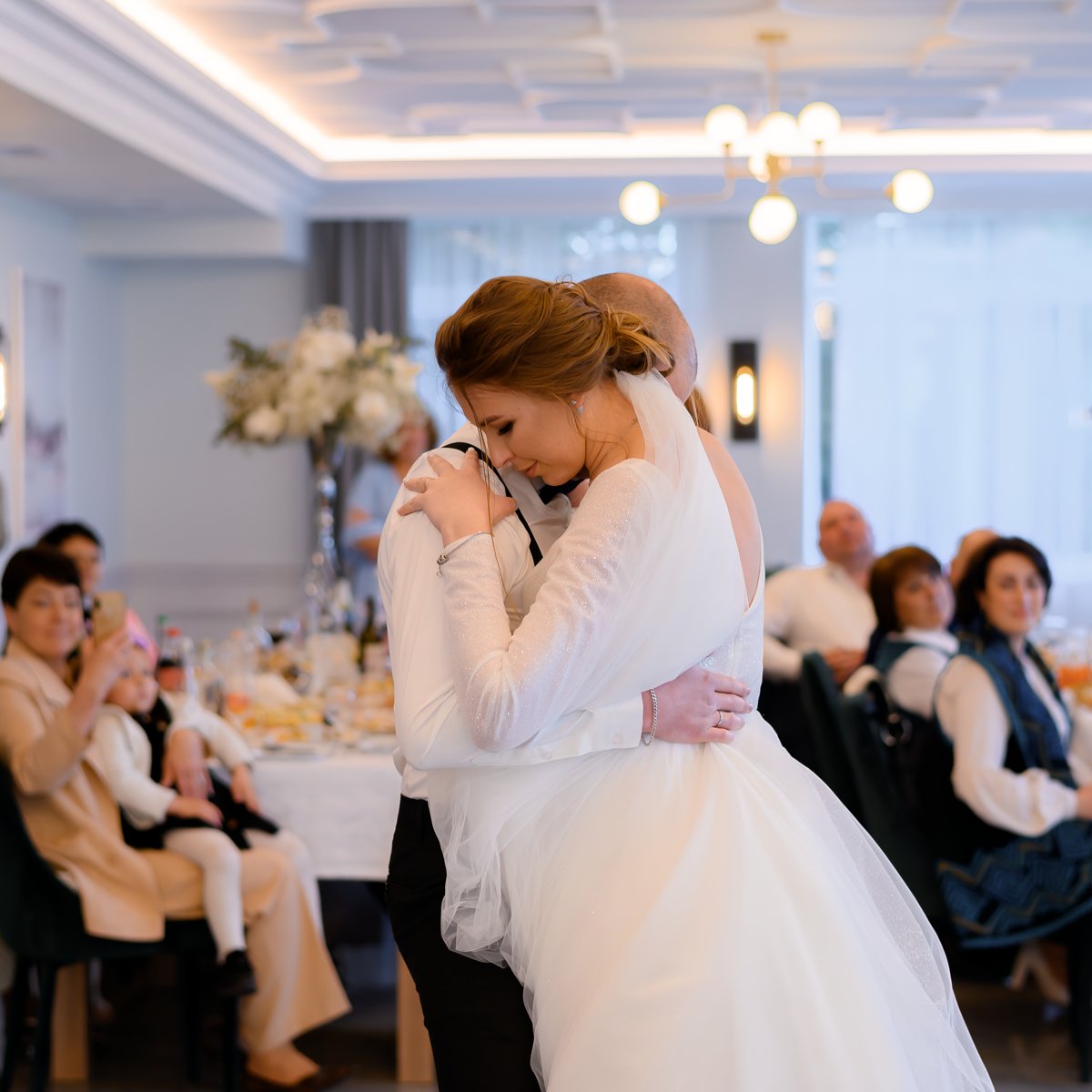 At their wedding reception, the bride and groom share an intimate embrace on the dance floor as guests look on from nearby tables.