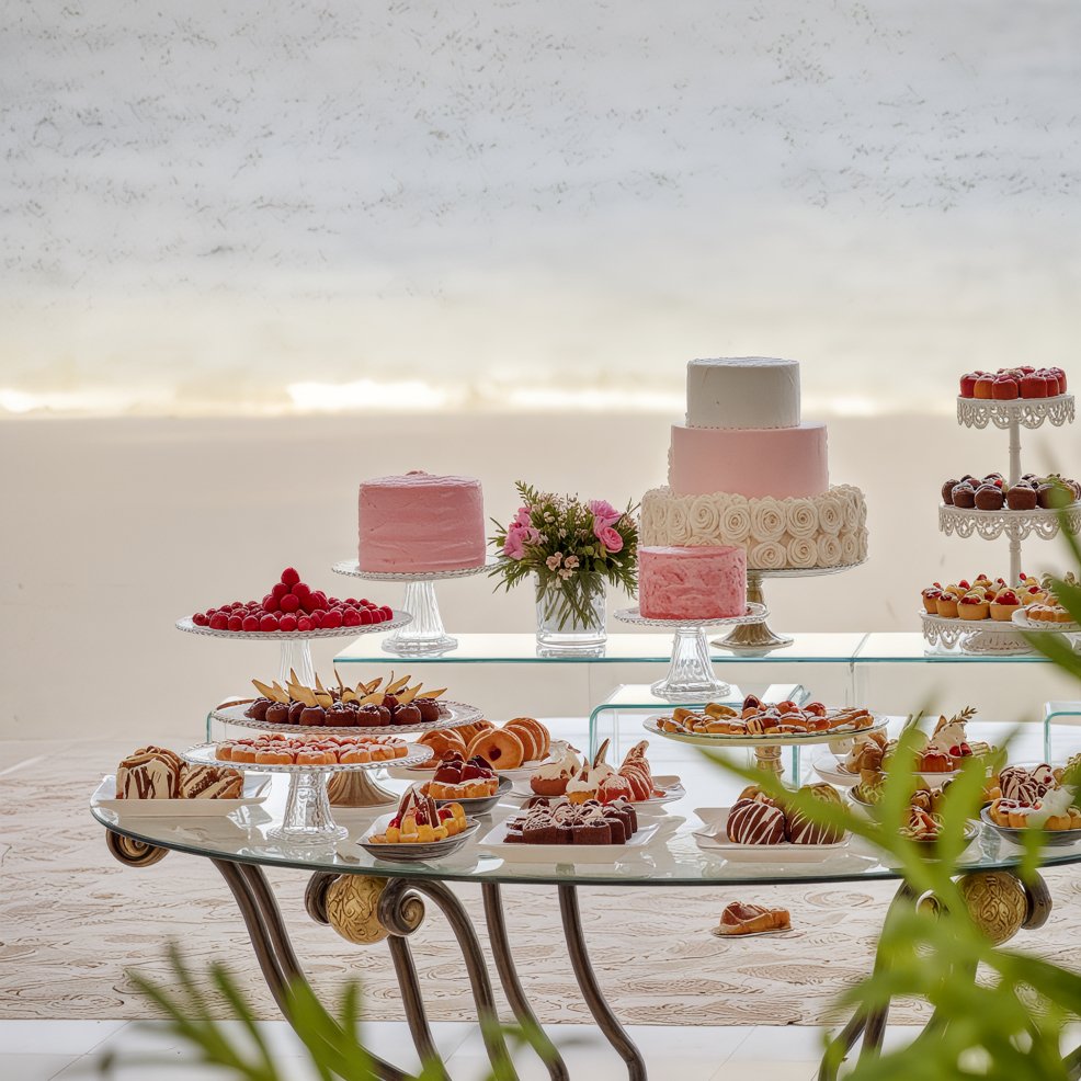 Table at a destination wedding with assorted pastries, cakes, and desserts on elegant glass stands.