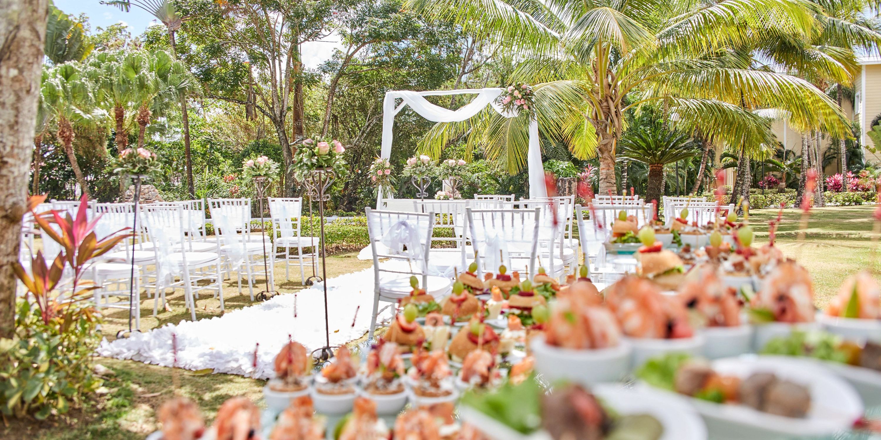 Bahia Principe Grand El Portillo: destination wedding outdoor setup with white chairs, floral arch, appetizers.