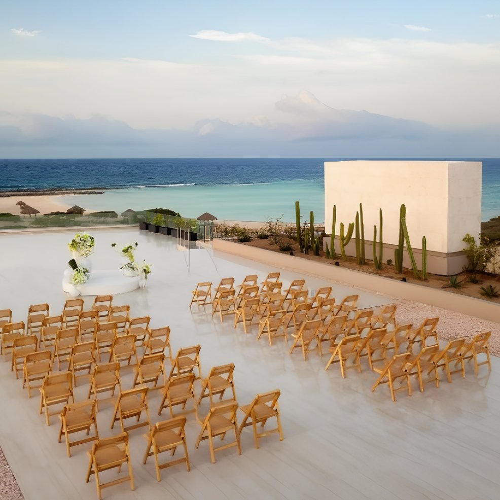 Rows of wooden chairs are arranged for a destination wedding, facing flowers by the ocean on a terrace.