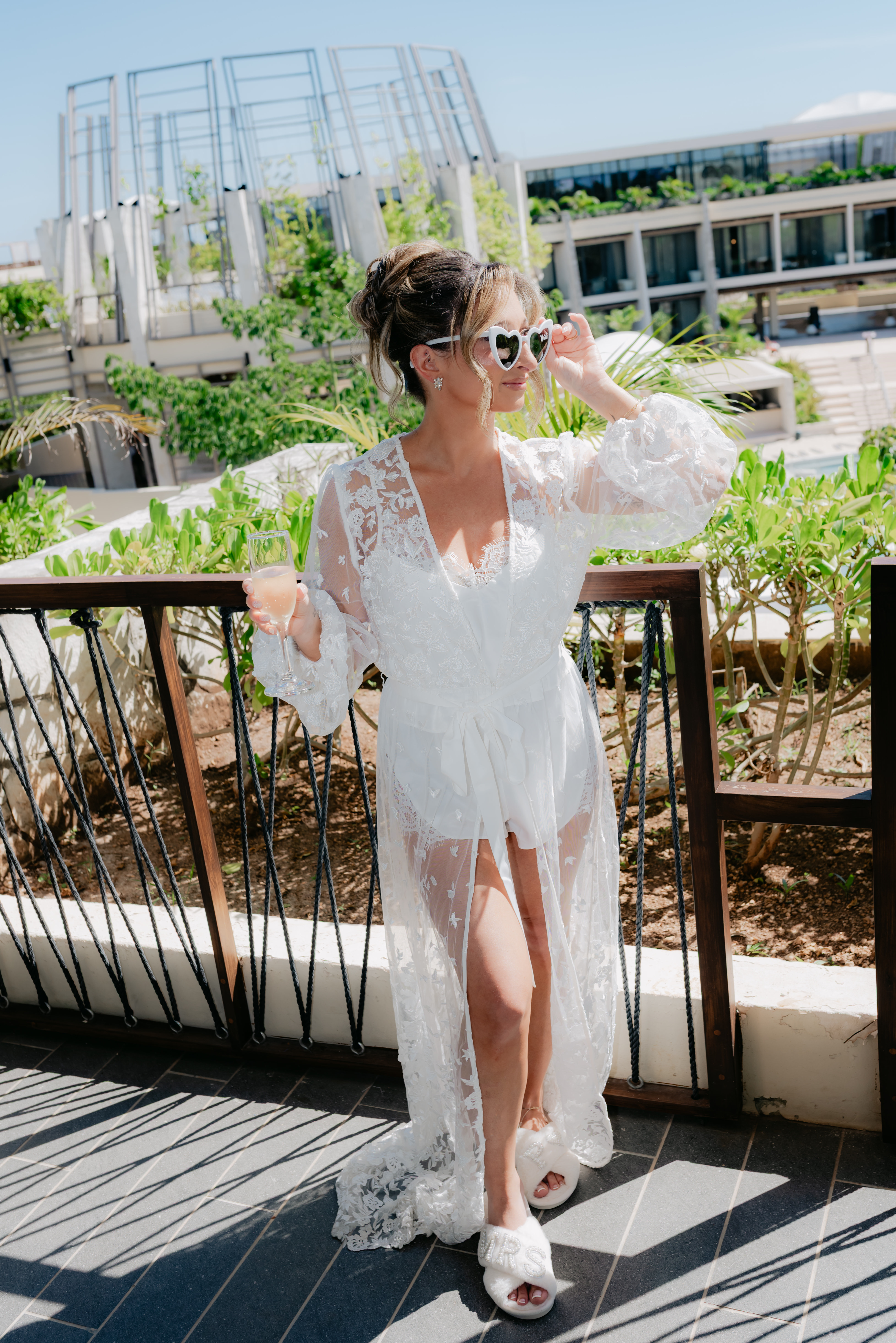 Woman in a Shirley and William (Copy) white lace robe stands on a balcony at a destination wedding, holding a drink.