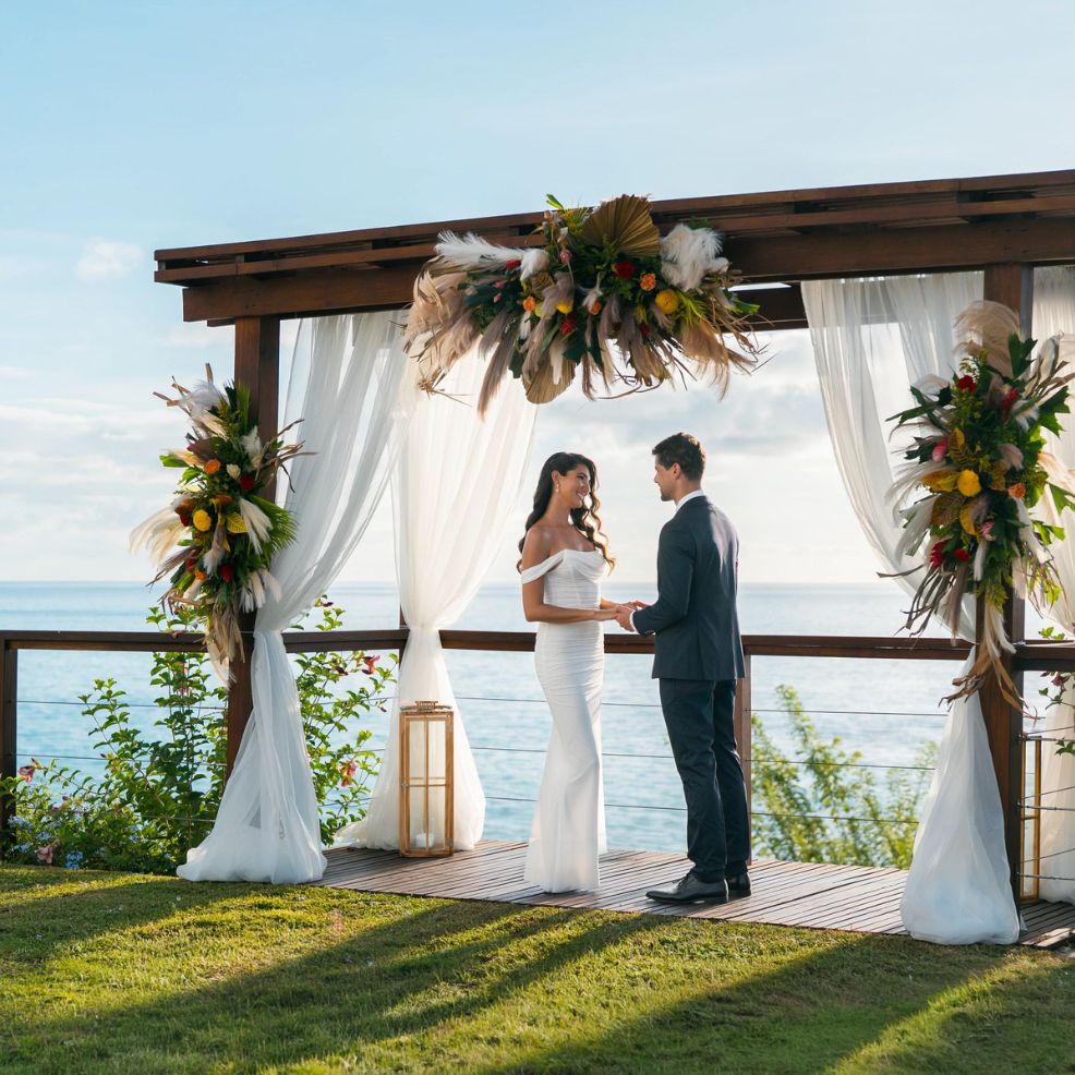 A couple holds hands under a decorated wooden arch by the sea during a destination wedding ceremony.