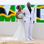 Bride and groom pose by a big "JAMAICA" sign at their oceanfront destination wedding.