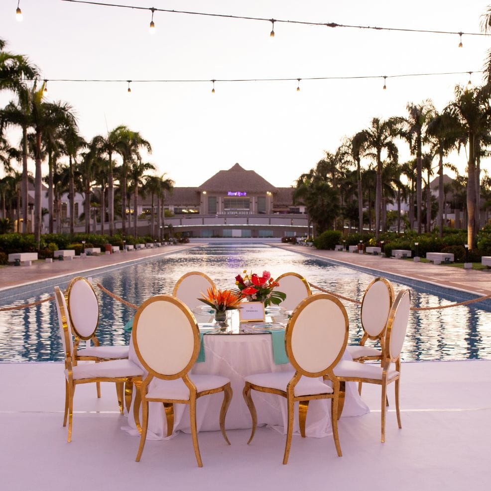 A round table with six chairs is set by a pool, perfect for a destination wedding at sunset amid palms.