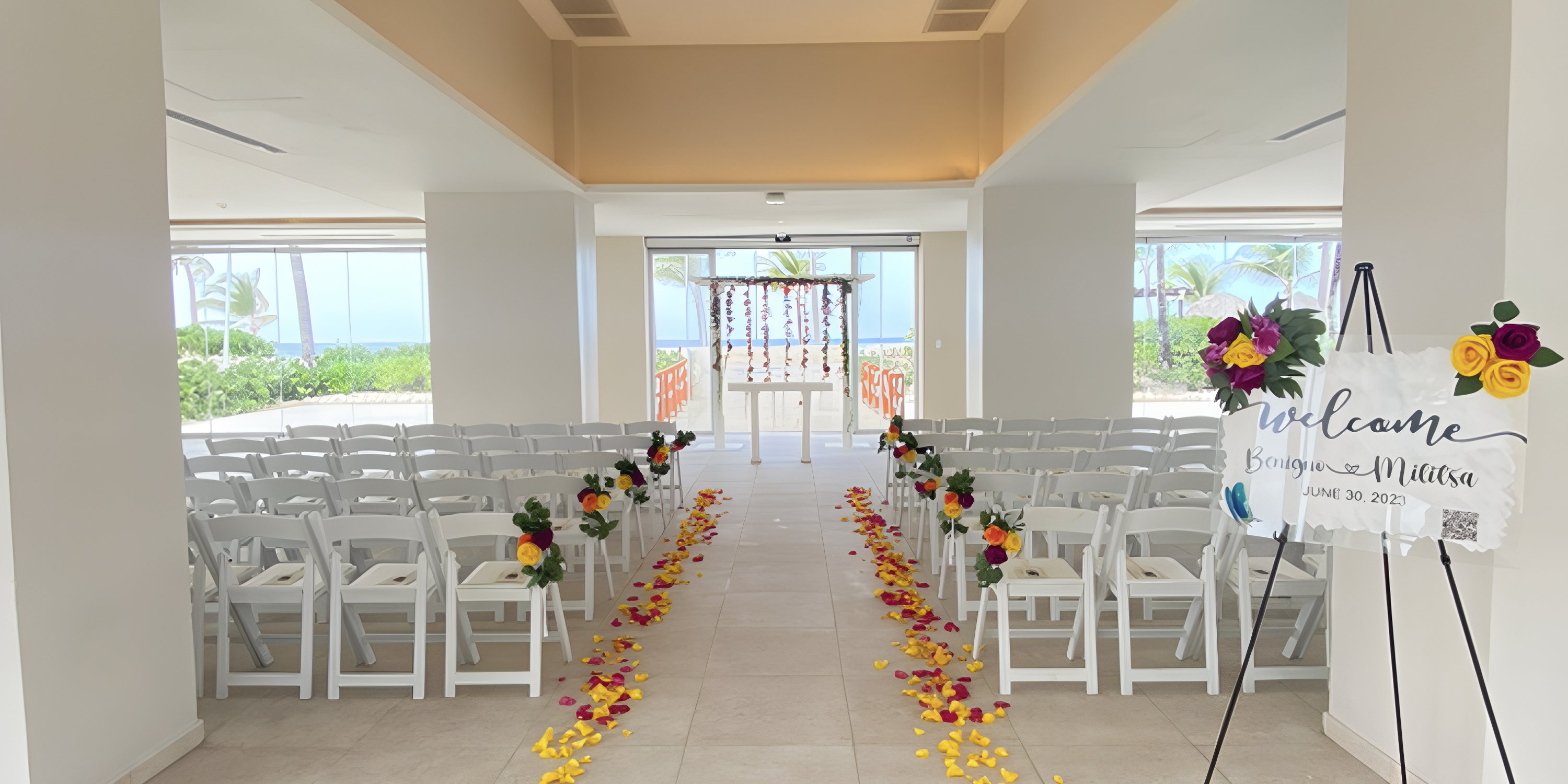 Destination wedding ceremony setup indoors with white chairs, aisle covered in petals, and a welcome sign.