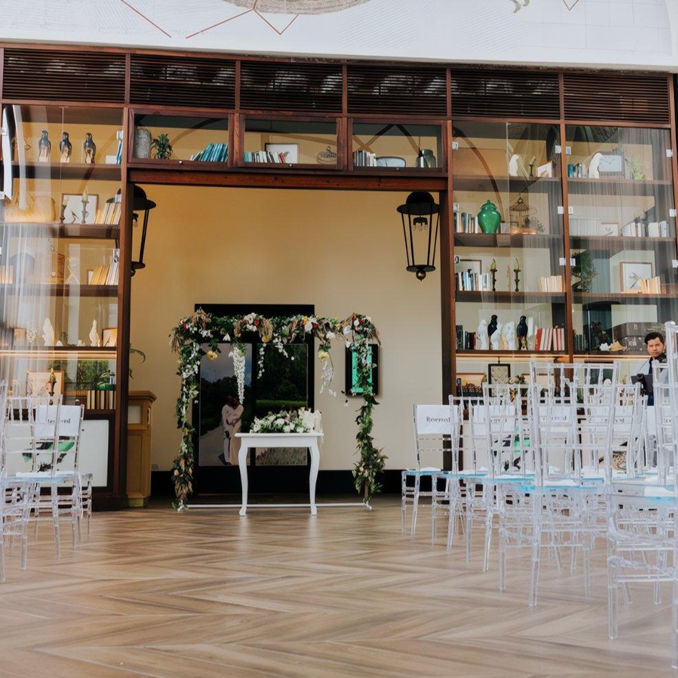 Destination wedding ceremony setup indoors with white chairs, floral arch, and decorative shelves behind.