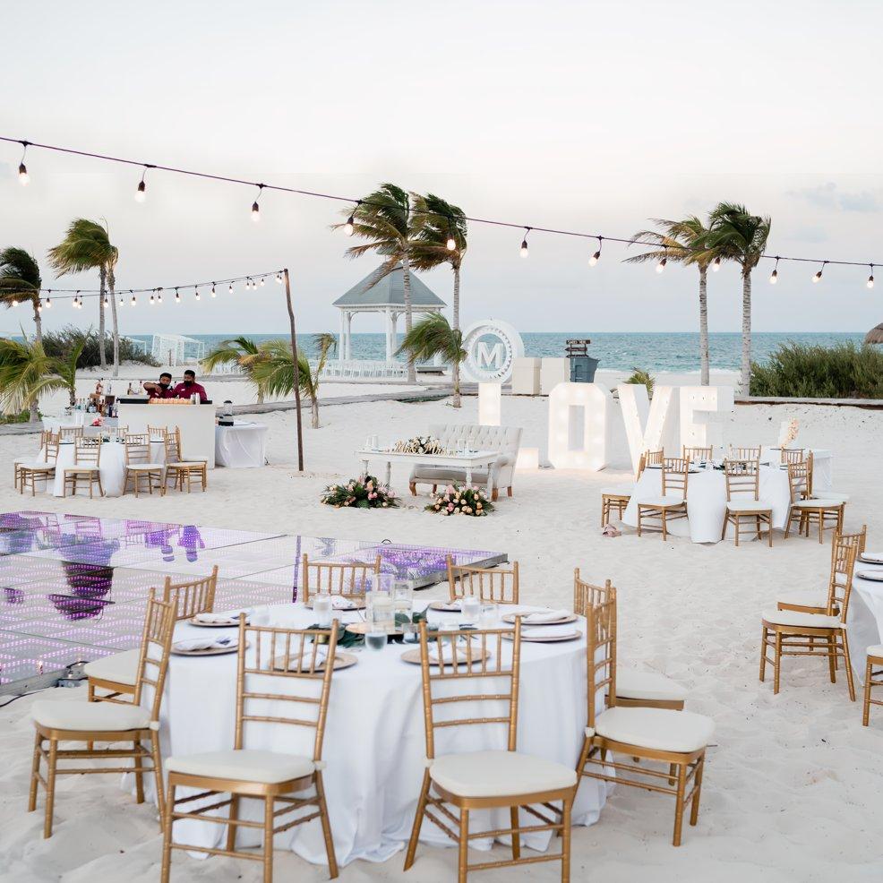 Destination wedding setup on the beach with round tables, string lights, and big "LOVE" letters by the sea.