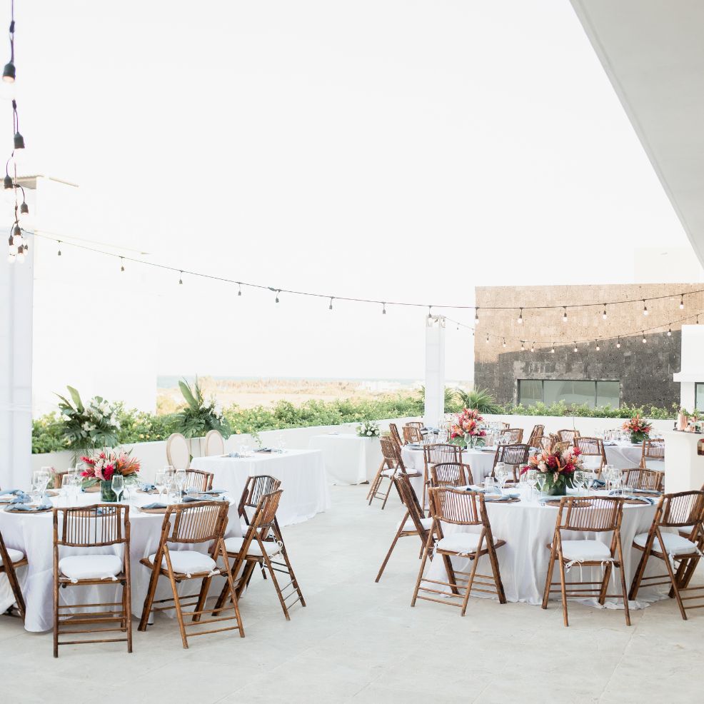 Destination wedding venue featuring round tables, wooden chairs, floral centerpieces, and string lights.