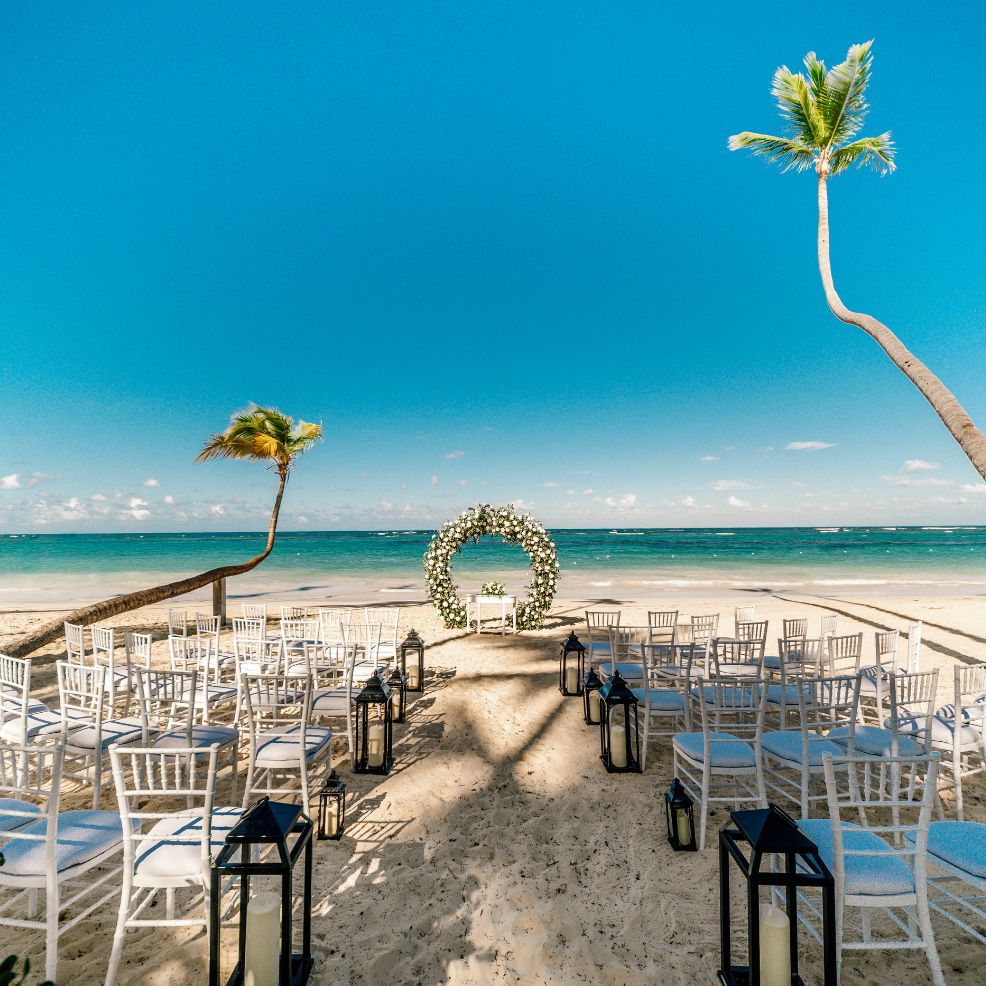 White chairs and lanterns set up on a sandy beach for a destination wedding facing a floral oceanfront arch.