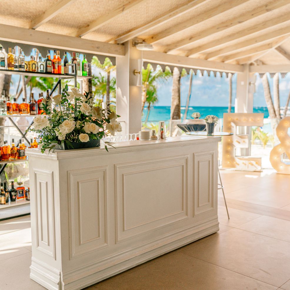 White bar counter with floral décor, liquor shelves, and a beach view perfect for a destination wedding.