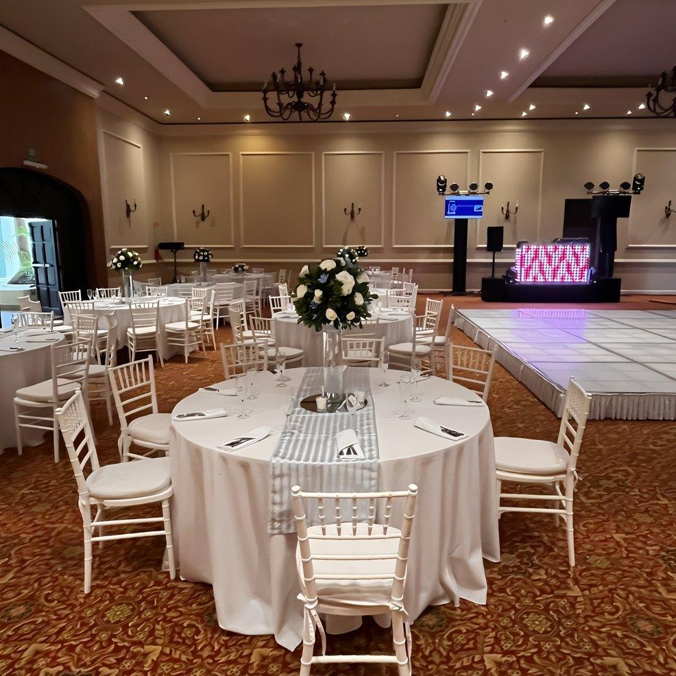 Round tables with white chairs and floral centerpieces set for a destination wedding in an event hall.
