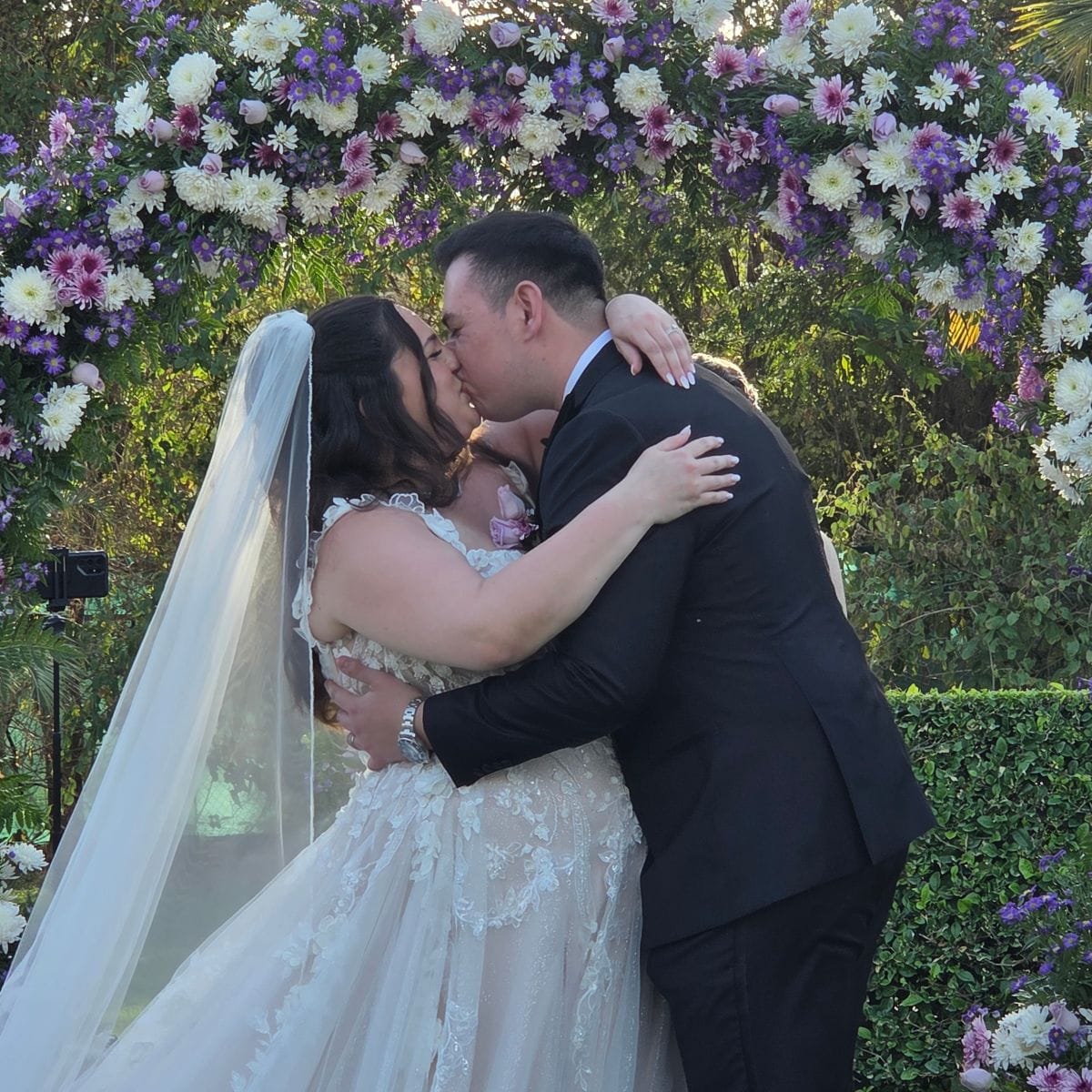 Ashley and Zach kiss beneath a floral arch during their romantic outdoor destination wedding ceremony.