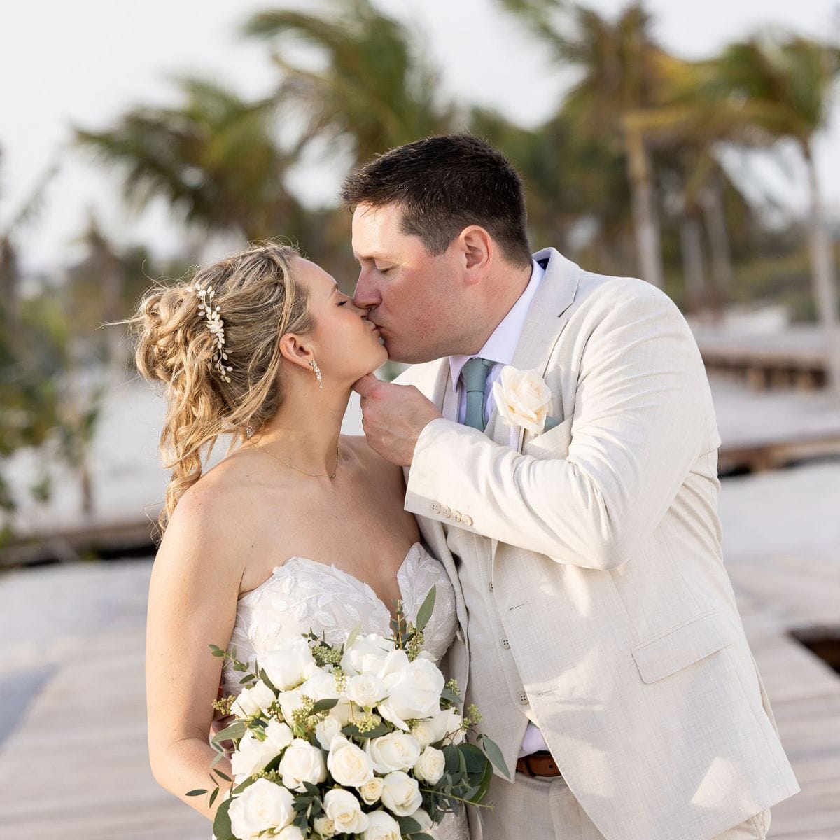 Lauren and Ben share a kiss at their destination wedding, holding a bouquet of white roses outdoors.