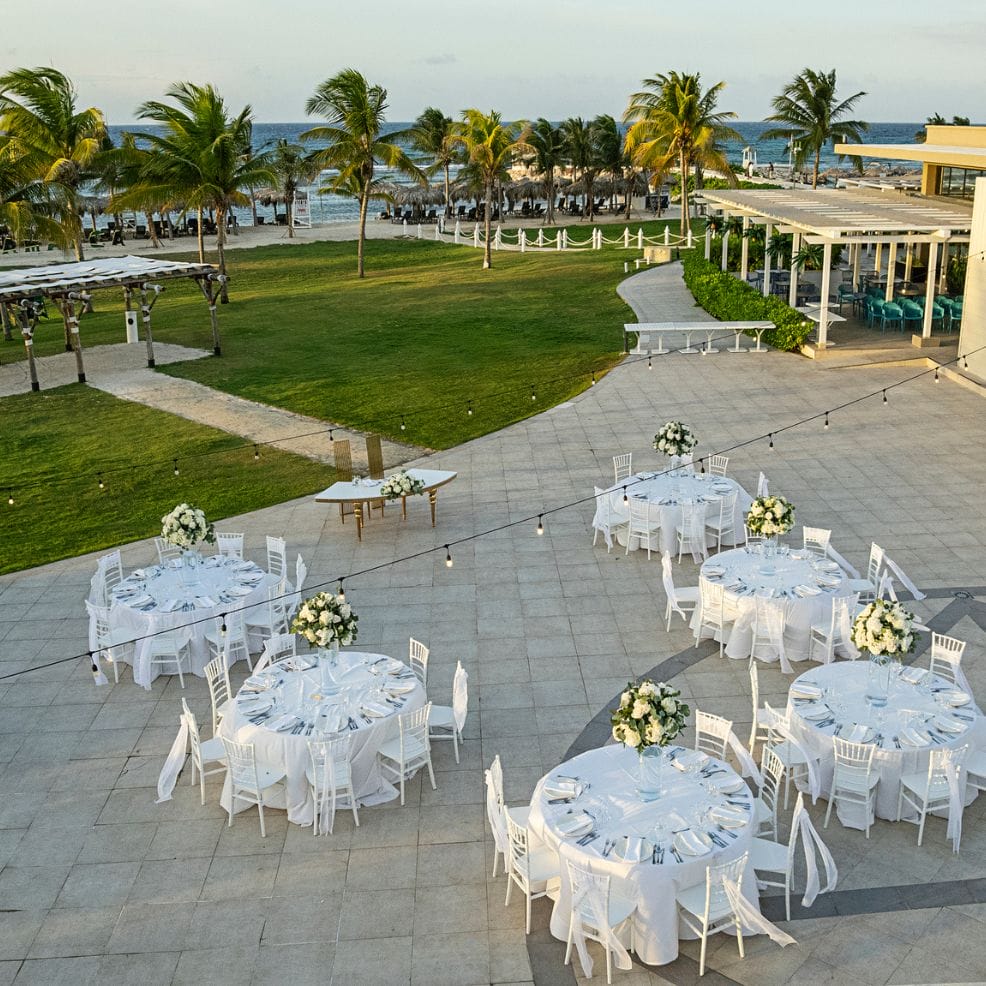 Round tables with white chairs arranged for a destination wedding by the beach with palm trees.