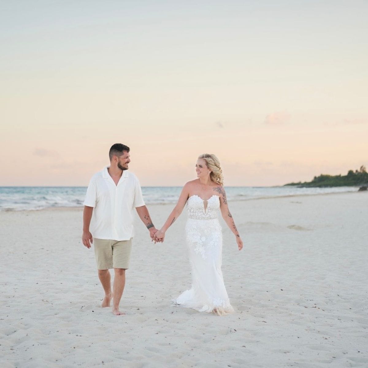 Hailey and Spencer stroll hand in hand along the beach at sunset during their destination wedding.