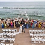 A destination wedding group stands on a deck by the ocean, empty chairs visible in the foreground.