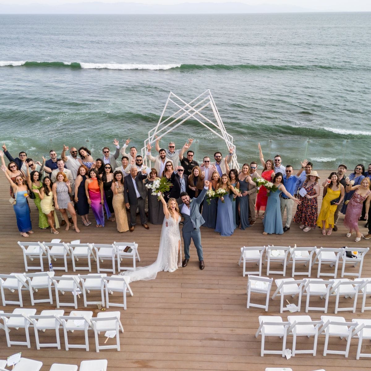A destination wedding group stands on a deck by the ocean, empty chairs visible in the foreground.