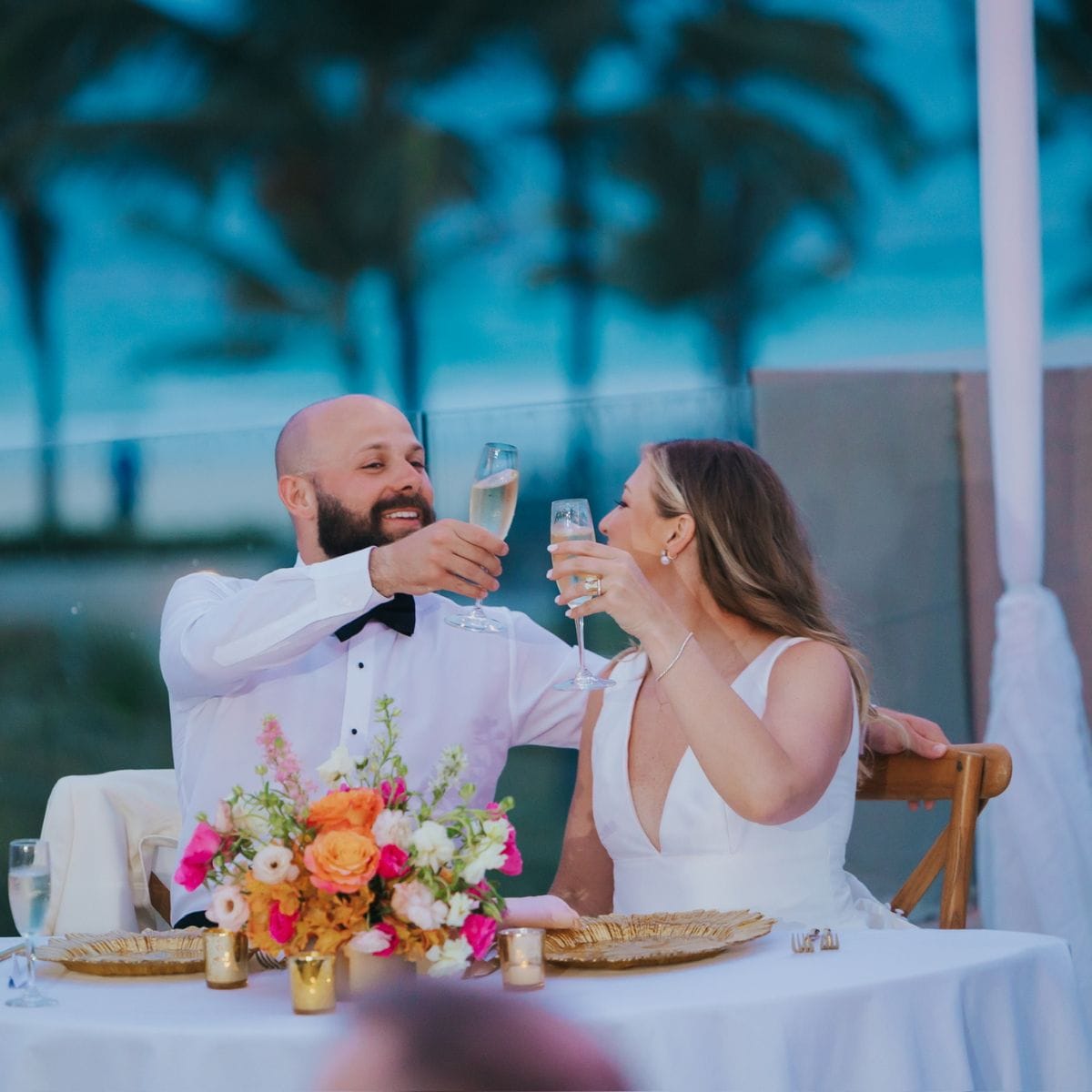 A bride and groom toast with champagne at a decorated outdoor table during their destination wedding.