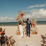A couple celebrates their destination wedding by walking under a floral arch on the sandy beach.