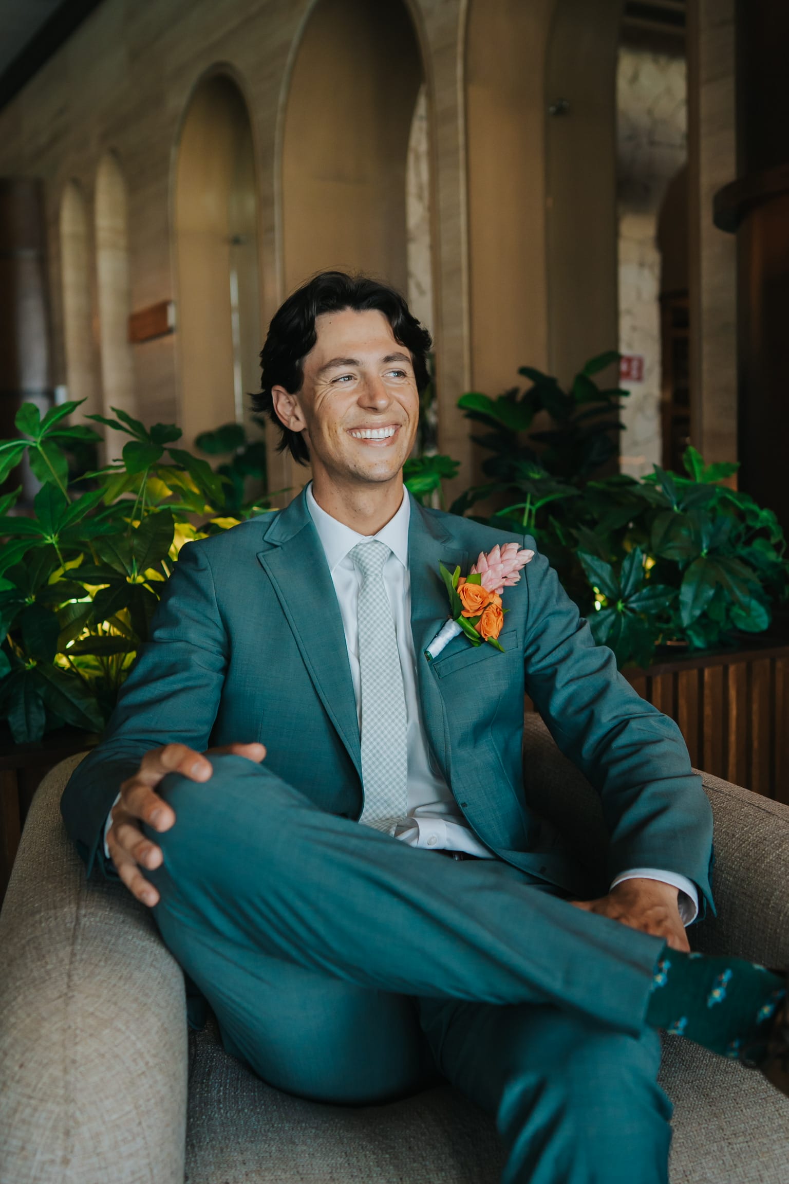 Man in Madison and Blake teal suit with orange boutonniere smiles at a destination wedding indoors.